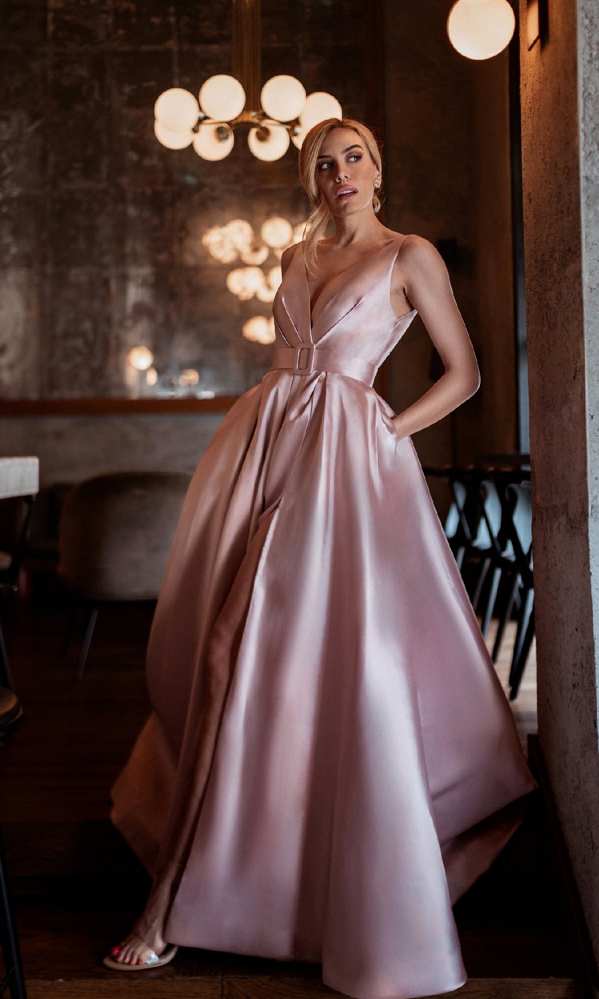 A woman wearing the Tina Holly BB209 Belted Long Prom Ball Gown, a floor-length, light pink satin A-line dress with a deep V-neckline, stands indoors near a concrete wall and modern light fixtures.